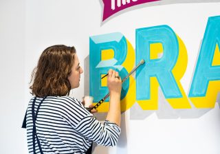 This World Belongs to the Brave A woman hand-painting lettering on a wall