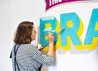 This World Belongs to the Brave A woman hand-painting lettering on a wall