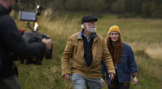 man shooting with camera as actors walk through a field