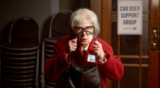 closeup photo of character, Betty, who is an older woman with black glasses and a red coat