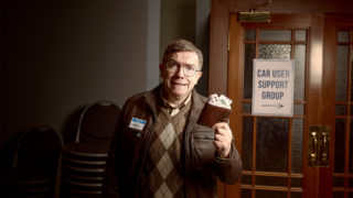 closeup photo of character, Neil, who is an older man with glasses and a plaid sweater vest