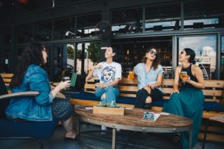 four friends drinking beer on a bench with a table in front