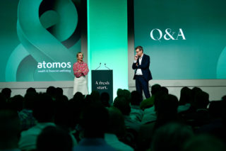 man and woman speaking on a stage with a green background and a large group of people watching