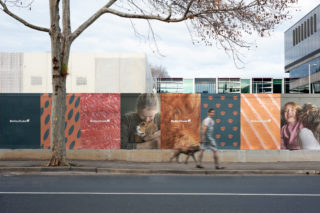 a photo of a man with his dog walking down a sidewalk with bella & duke ads lining a wall which he's walking in front of