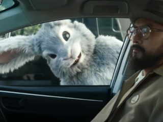 A super bowl commercial still of a bunny peering in through the passenger side window of a car at a man with a dark beard, glasses, and hat