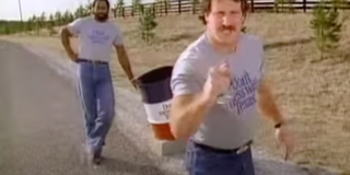 Original man in a t-shirt with a moustache points at the camera while a man behind him leans against a trash bin