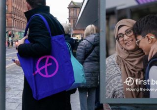photo of a man standing at a bus stop with a purple and pink first bus tote