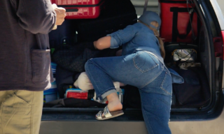 Colorectal Cancer Campaign - Take a Look at Your Trunk still frame of a woman with long hair and jeans facing away from the camera digging through the trunk of a car with a man standing and watching