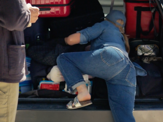 still frame of a woman with long hair and jeans facing away from the camera digging through the trunk of a car with a man standing and watching