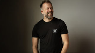 a photo of andrew dobbie, ceo of madebrave wearing a black shirt and standing in front of a cream wall