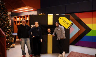 Refuweegee Winter Wrap-Up photo of four people smiling and standing in front of a giant present wrapped in black paper with a golden bow