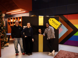 photo of four people smiling and standing in front of a giant present wrapped in black paper with a golden bow