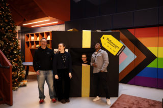 photo of four people smiling and standing in front of a giant present wrapped in black paper with a golden bow