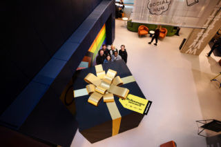 photo of six people from above. They're smiling and standing in front of a giant present wrapped in black paper with a golden bow