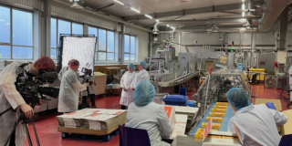 Tunnock's Brand Film - Behind the Scenes a photo of two tunnocks employees being filmed by a man with a camera. Setting is inside the tunnocks factory with other tunnock's employees watching from behind the scenes